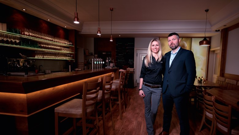Moravian Embassy, © tonality Two people stand in a stylish bar with a wooden counter and glass shelves.