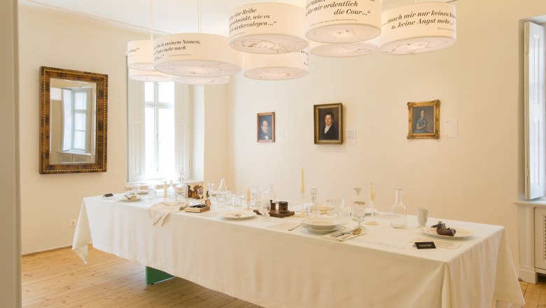 Beethoven House table, © Stadtgemeinde Baden A laid table in a room with portraits on the wall and hanging lamps with texts.