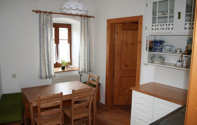 Kitchen, © Grünfelder Small kitchen with wooden furniture, dining table, chairs and a window with curtains.