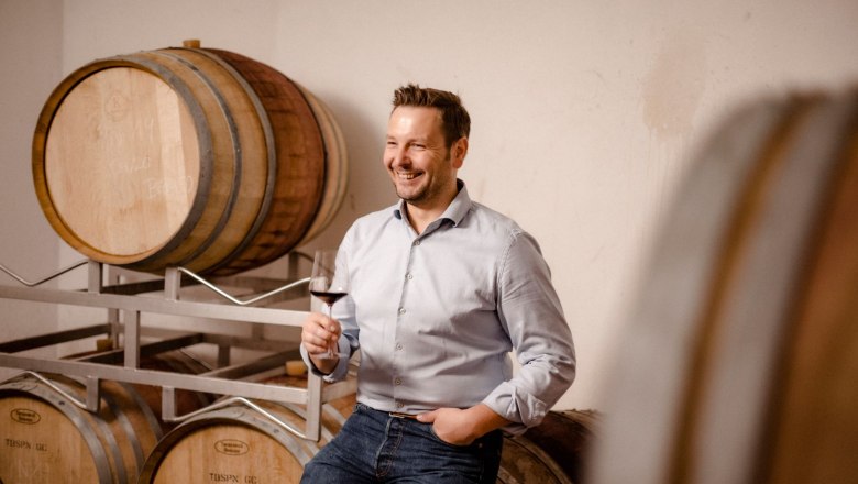 Heinisch Winery, © Astrid Bartl A man leans smiling against wine barrels and holds a glass of red wine in his hand.