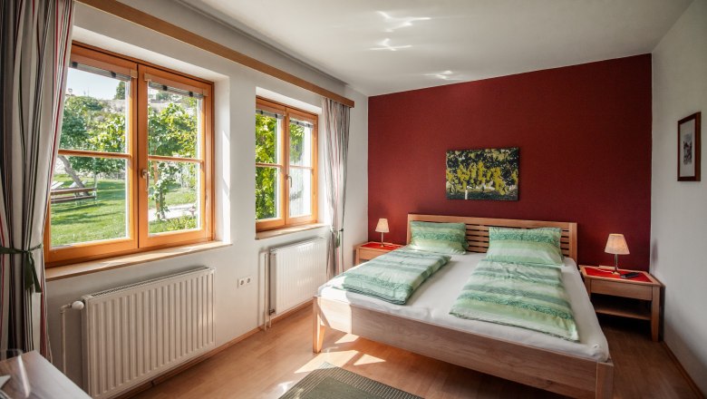 Guesthouse-Winzerhof Bernhard, © Jolly Schwarz Bright bedroom with double bed, red walls and large windows with a view of the greenery.
