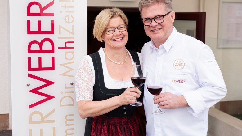 Take your time - Restaurant Der Weber, © Hotel Weber/Giefing A man and a woman clink glasses of wine next to a sign that reads 'The Weaver'.