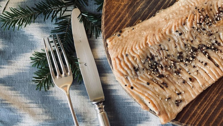 Smoked carp, © Amelie Seilern-Aspang Close-up of a smoked carp fillet on a wooden plate, decorated with peppercorns. Next to it are a fork and a knife on a blue cloth.
