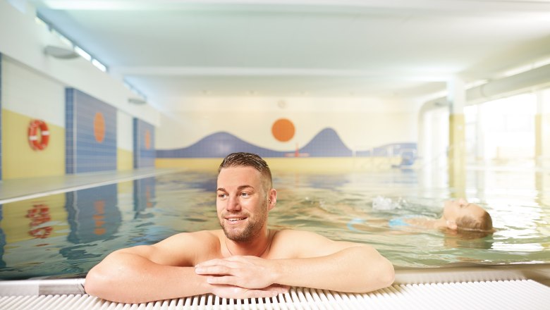 Zwentendorf indoor swimming pool, © Florian Schulte A man leans against the edge of an indoor swimming pool while a person swims in the water in the background.