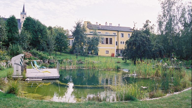 Swimming biotope in the large natural garden, © Ludwig Schneider A swimming pool in a large garden with a yellow building in the background.