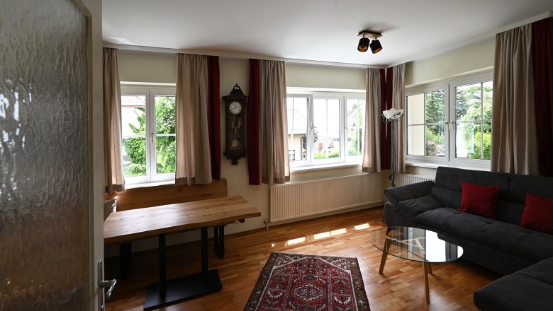 Living room, © Christine Kuppelwieser A living room with wooden floor, gray sofa, glass table, wall clock and carpet. Large windows with curtains let in plenty of light.