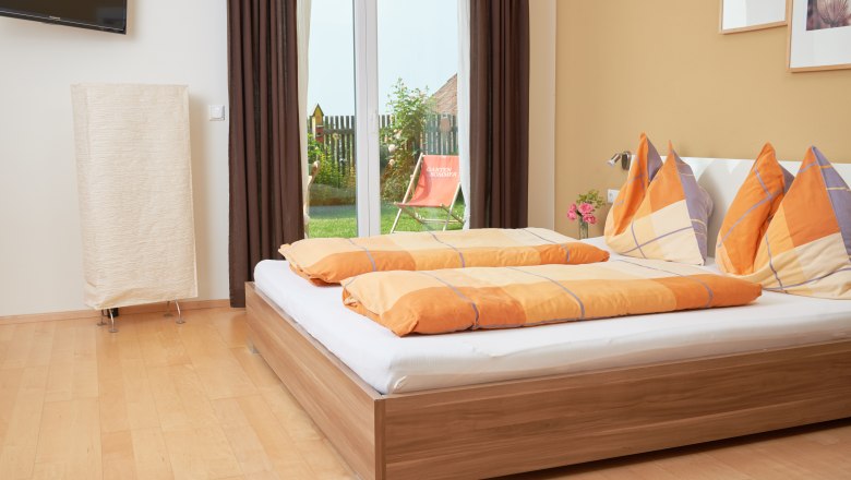 Wood hay room, © Philipp Monihart A cozy bedroom with a double bed, orange comforters and a view of a garden through a glass door.