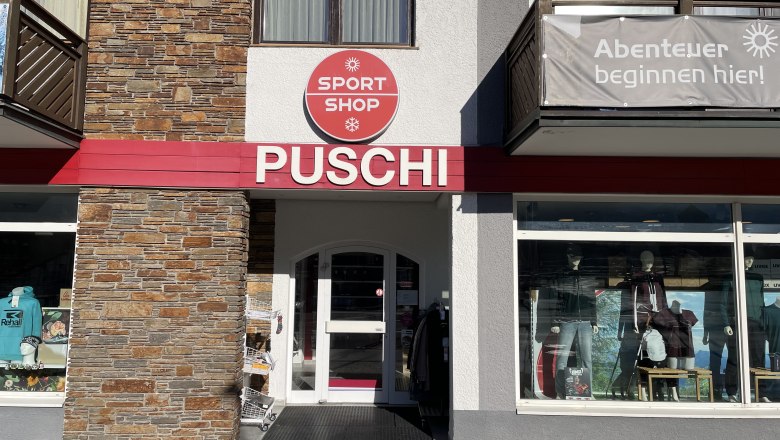 Sport-Puschi-Sportshop-Front, © Sport Puschi Entrance area of a sports store with the inscription 'PUSCHI' and a sign 'SPORT SHOP'. Shop window with sportswear and equipment.