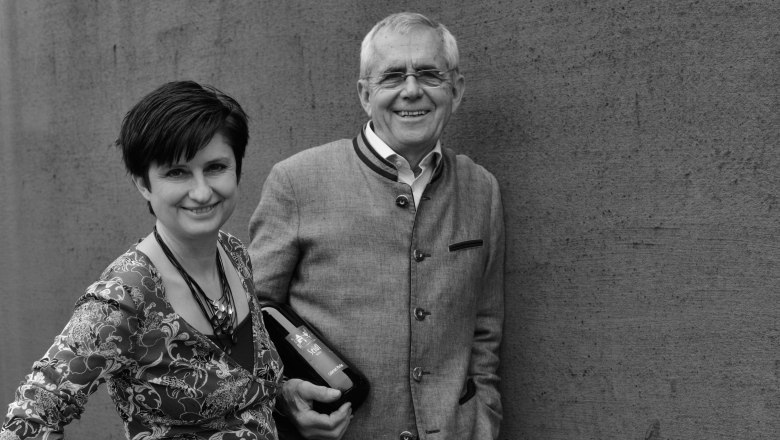 Andrea and Gerhard Seidl, Seidl Winery, Bruck/Leitha, © Die Rubin Carnuntum Weingüter A man and a woman stand smiling in front of a wall. The man is holding a bottle of wine.