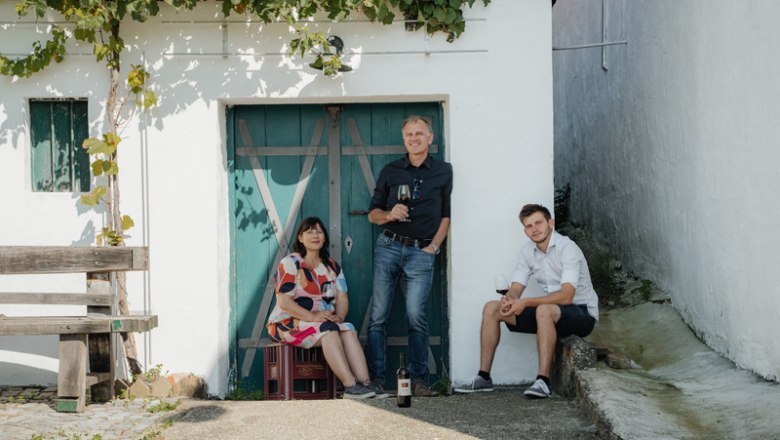 Family Bauer, © Martin Mattes Three people in front of a wine cellar with a blue door and grapevines.