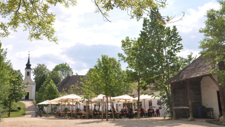 Village inn in the museum village, © Museumsdorf / Bodensteiner A village inn in the museum village with parasols and a church in the background.