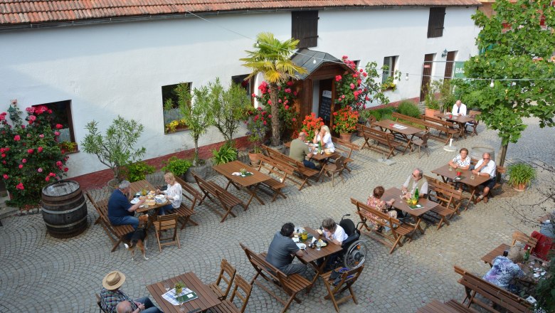 Wine tavern in the courtyard, © Sophie Kienberger People sit at wooden tables in a paved courtyard of a wine tavern, surrounded by flowers and trees.