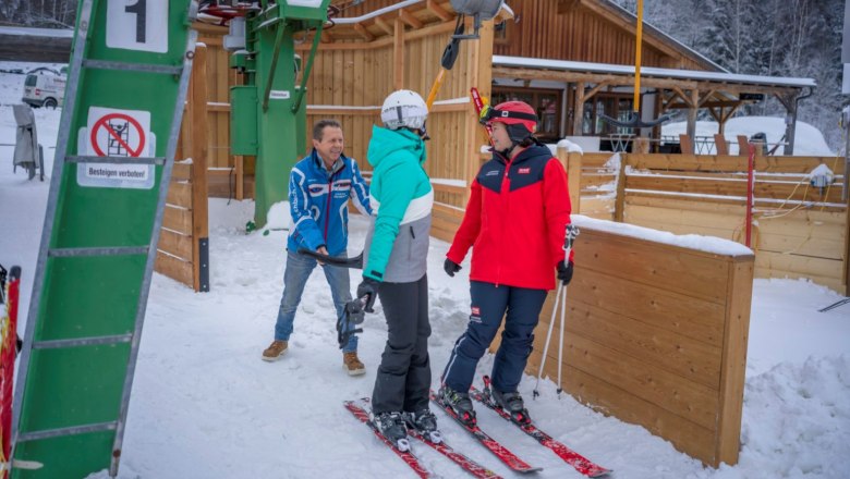 T-bar lift, © Waldviertel Tourismus, Robert Herbst Two female skiers stand at a ski lift in a snowy setting while a man helps them.