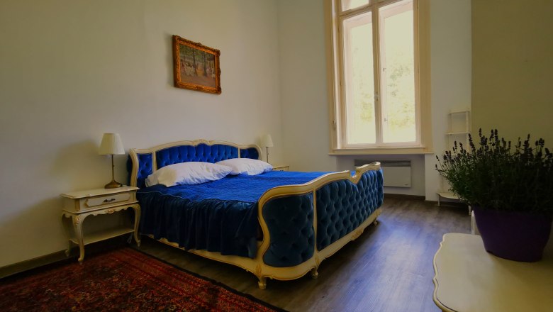 Bedroom in the Nikolaus apartment, © Herrnhof Elegant bedroom with blue bed, antique furniture and large window.