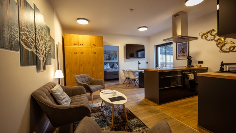 Ötscher Lodge, © Niederösterreich Werbung / Maximilian Pawlikowsky Modern living room with kitchen, sofa, armchairs and dining table. Light wood furniture and wall art can be seen.