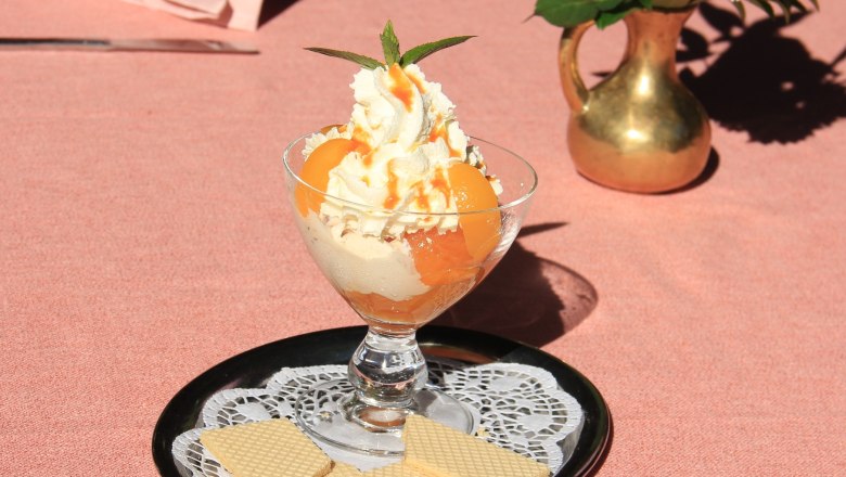 Apricot ice dream, © Schendl A dessert glass with ice cream, whipped cream and apricots on a tray with waffles, next to a vase with flowers.