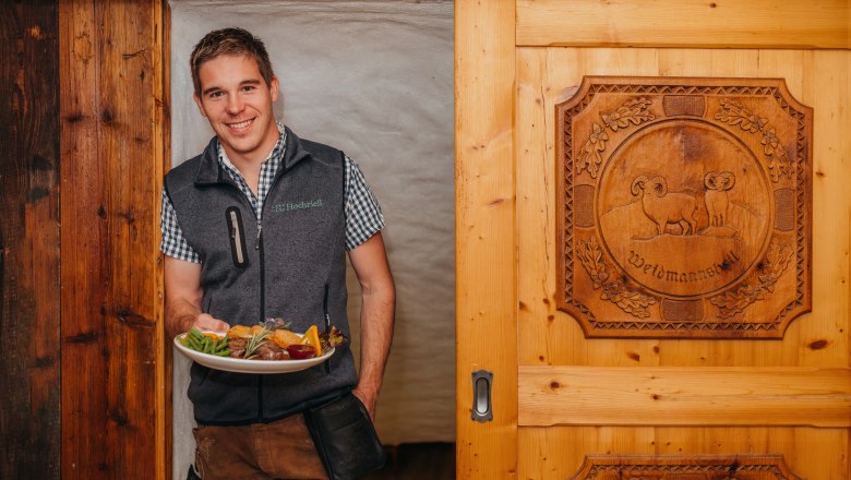In service: Landlord Matthias Distelberger, © Niederösterreich Werbung/Daniela Führer A man in traditional dress holds a plate of food in front of a wooden door with a carved motif.
