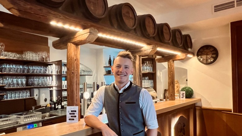 Landlord Matthias Bachofner, © Matthias Bachofner A man in traditional dress stands smiling at a bar with wooden barrels and glasses in the background.