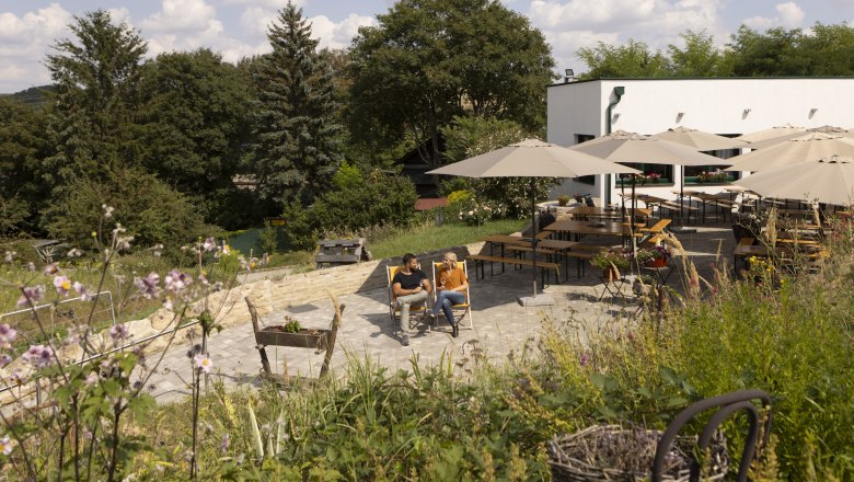 Winter wine tavern, © Weinviertel Tourismus GmbH / Schwarz-König Two people sit on deckchairs in a garden with parasols and tables, surrounded by trees and flowers.