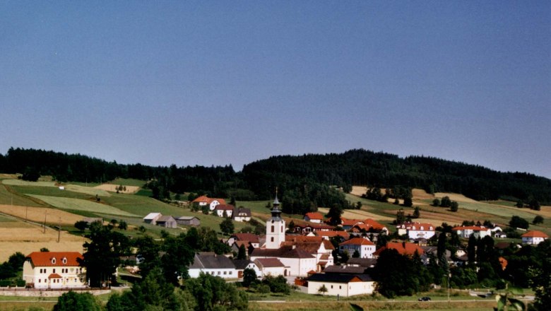 St. Martin, © Marktgemeinde St. Martin Panorama of a village with a church and red roofs, surrounded by green fields and forests.