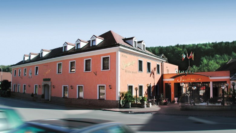 House, © Das Steinberger A pink-colored hotel building with several windows and an entrance with a canopy. A car drives past in the foreground.