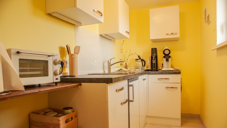 Kitchen, © Fam. Heinrich Small kitchen with yellow walls, white cupboards and appliances such as a toaster and coffee machine.