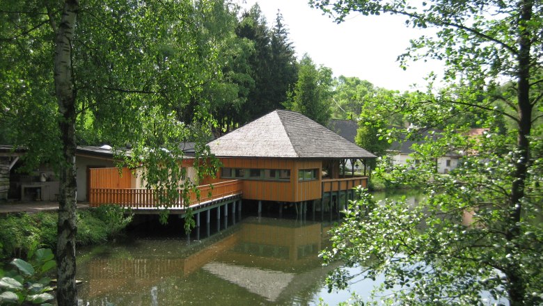 Waldviertler Forellenhof, © Gemeinde Jaidhof A wooden building on stilts above a pond, surrounded by trees.