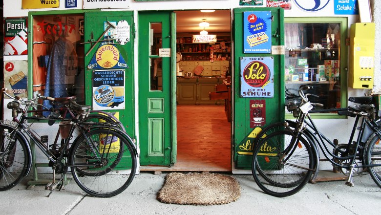 Museum, © Fam. Buchinger Entrance to an old store with a green door, surrounded by retro advertising signs and two bicycles.