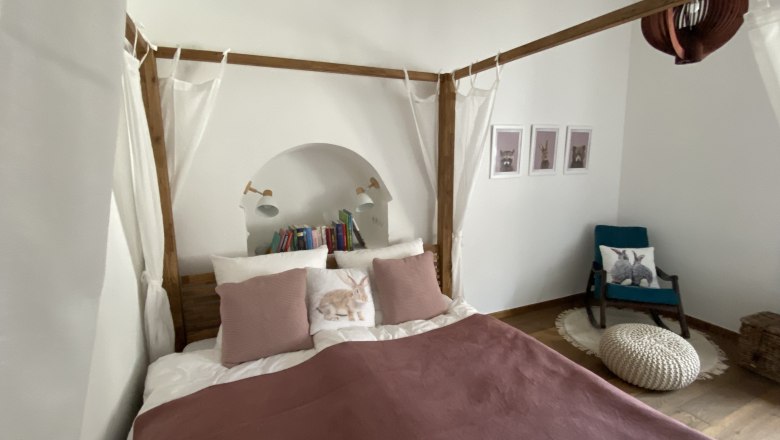 2nd bedroom, © Kathrin Bürger Cozy bedroom with four-poster bed, pillows, books and an armchair with rabbit cushions.