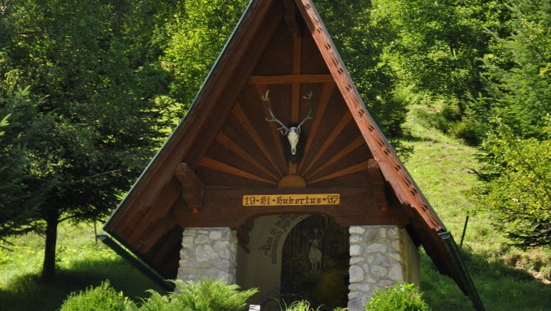 Hubertus Chapel, © Schwarzenbach/Piel. A small chapel with a pointed roof and deer antlers, surrounded by trees and bushes.