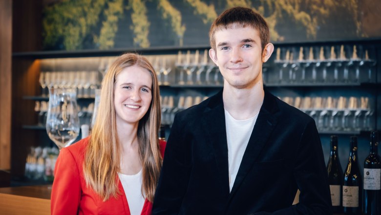Katharina and Lorenz Baumgartner, © M. Ledwinka Katharina and Lorenz Baumgartner in elegant clothes in front of a shelf with wine glasses.