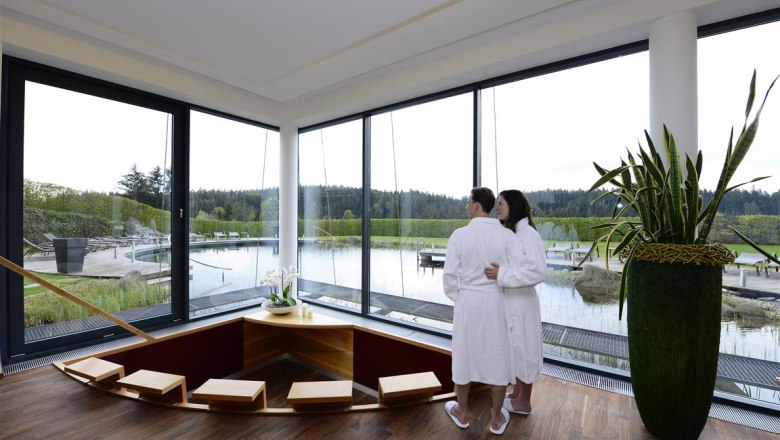 time out_wellness_04, © Hotel Schwarz Alm A couple in bathrobes stand in a modern wellness area with a view of a pond.
