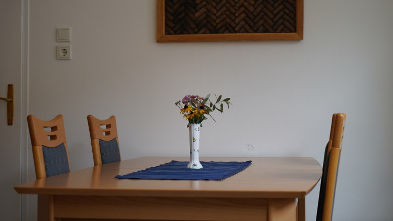 Country house Krell, © Weingut Krell A table with four chairs, a vase of flowers and a blue cloth in a room with a white wall and wall decor.