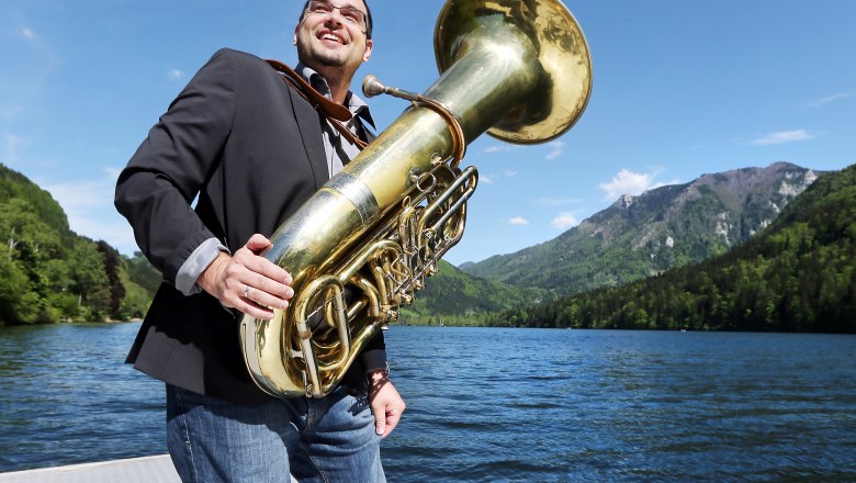 wellenklaenge: Art on the lake, © weinfranz.at A man with a tuba stands barefoot on a jetty by a lake, surrounded by mountains and blue sky.