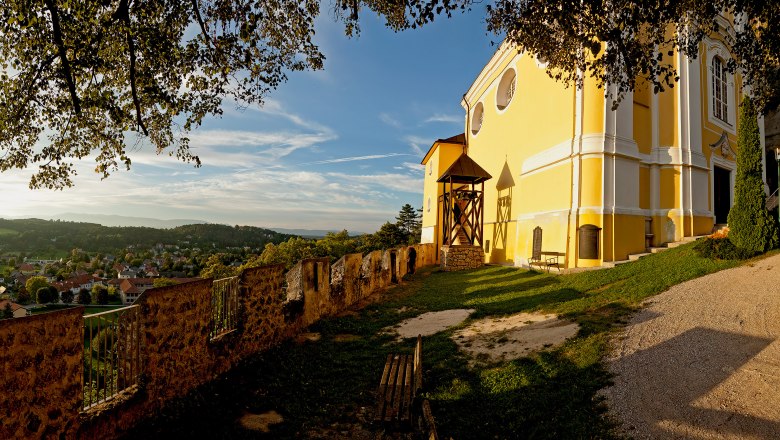 Viewpoint at the mountain church in Pitten, © Wiener Alpen, Franz Zwickl Yellow church on a hill with a view of a village and wooded hills in the background.
