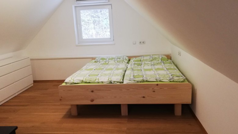 Double bed, © Günter Lammer An attic room with a wooden double bed and green bed linen, with a white chest of drawers next to it.