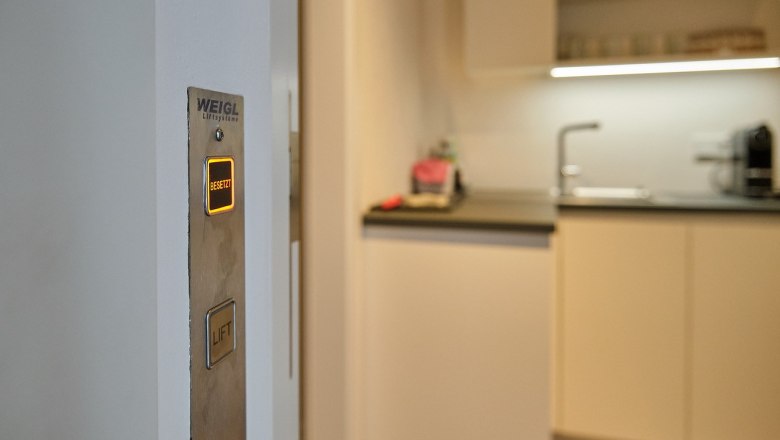 Elevator directly to the living areas, © Prof. Arch. DI Dietmar Kraus An elevator button with the inscription 'Busy' lights up in a modern living room next to a kitchen.