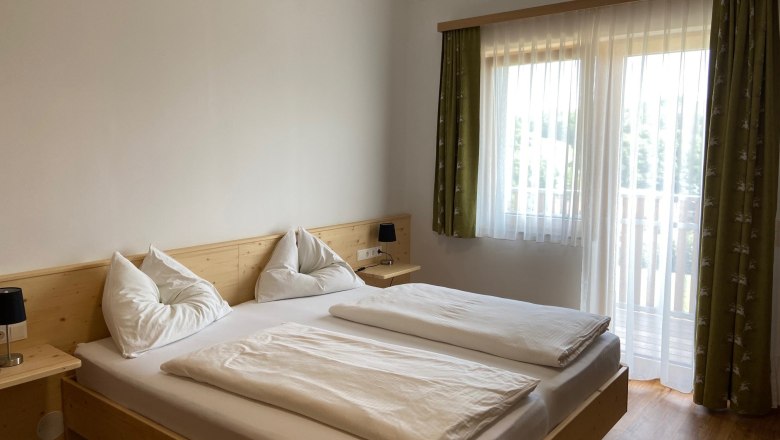 Family room, © Wiener Alpen A bright bedroom with a double bed, white comforters and pillows. A window with green curtains lets in natural light.