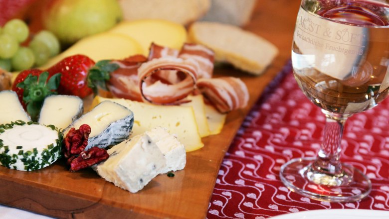Brettljause, © weinfranz.at A snack with cheese, ham, bread and fruit on a wooden board, with a glass of white wine next to it.