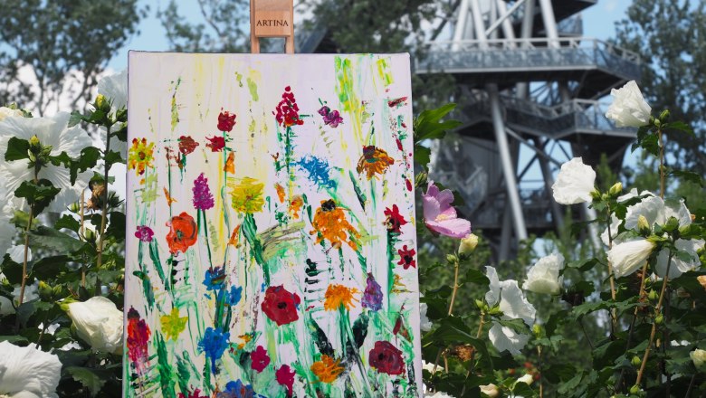 Flower meadow GARTEN TULLN, © Art Addventure A painting of colorful flowers stands on an easel in a garden, surrounded by white blossoms. A lookout tower can be seen in the background.