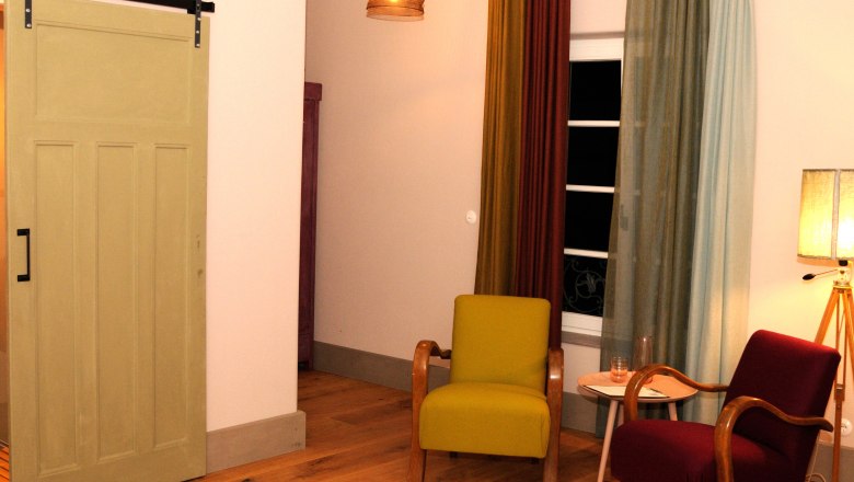 Room 5, © Franz Böck A cozy room with two armchairs, a small table and a lamp. A sliding door and colorful curtains can also be seen.