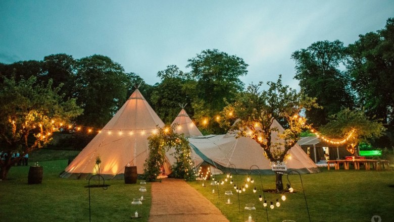 Tipi in the area, © Szilagyi An illuminated teepee tent in a garden with fairy lights and trees at dusk.
