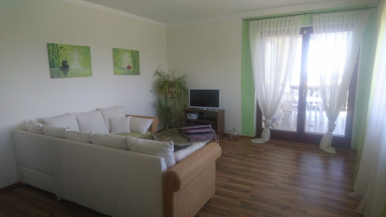 Living room, © Anke Spreitzer A living room with a white sofa, a small TV, plants and large windows with curtains.
