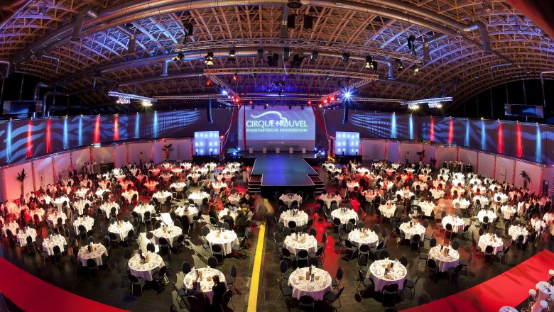 ARENA NOVA Gala, © ARENA NOVA Large event room with round tables and stage, illuminated in red and blue.