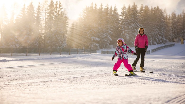 Family ski area at the Wexl Arena St. Corona am Wechsel, © Familienskiland der Wexl Arena St. Corona am Wechsel Child and adult skiing in the snow, surrounded by trees and sunshine.