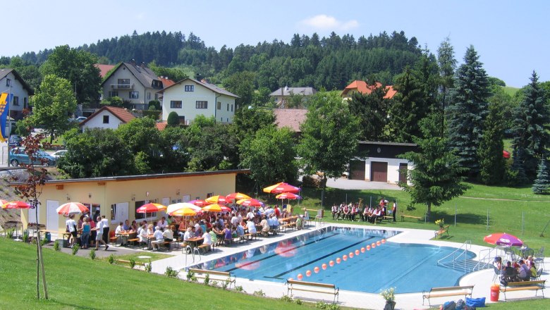 Outdoor pool Els 01, © Ingrid Kleber Outdoor pool with swimming pool, sunshades and people on deckchairs, surrounded by green hills and houses.