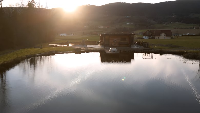 View of the pond, © Familie Moser A pond with a wooden building on the shore, surrounded by hills and trees at sunset.