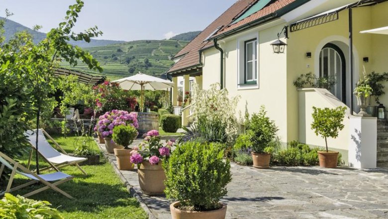 Country house Smöch, © Familie Smoech An idyllic country house with a well-tended garden, flowering plants and sun loungers under a sunny sky.