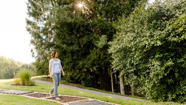 In the spa gardens of Bad Schönau, © Niederösterreich Werbung/Doris Schwarz-König A woman walks barefoot on a green path in the spa gardens of Bad Schönau, surrounded by trees and sunlight.