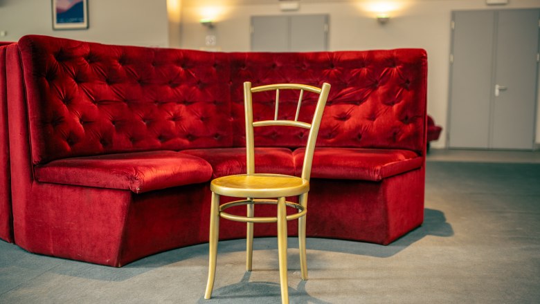 Reichenau_Chair in the theater, © wieneralpen_fueloep A single wooden chair stands in front of a red velvet bench in a theater foyer.